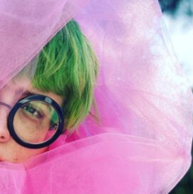 Person with green hair and large black framed glasses peering out of pink tulle